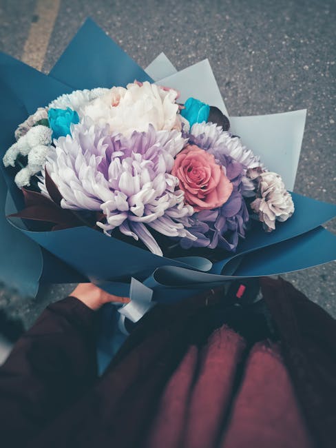 A hand holding a vibrant floral bouquet wrapped in dark blue and light blue paper, featuring a mix of fresh flowers including large white and purple chrysanthemums, a pink rose, and blue accents. The 