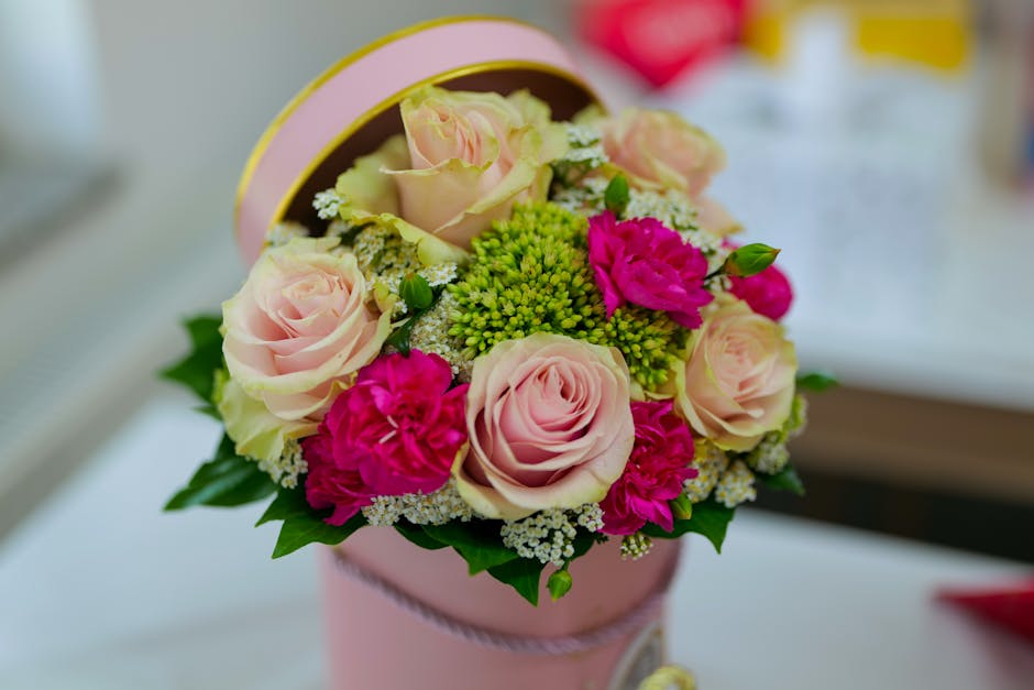 A close-up of a floral gift arrangement featuring soft pink roses, vibrant fuchsia carnations, and green filler flowers arranged in a round, pastel pink gift box with a delicate ribbon. The bouquet is