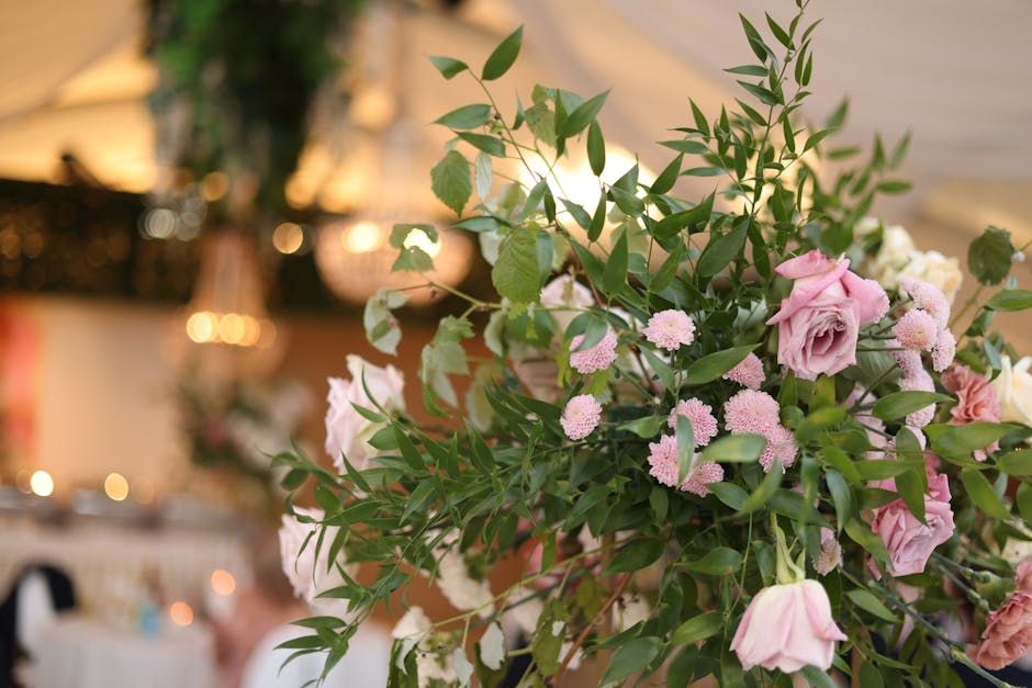 A lush floral arrangement featuring soft pink roses, small pink chrysanthemums, and delicate white blossoms, complemented by an abundance of green foliage with elongated, pointed leaves. The bouquet i