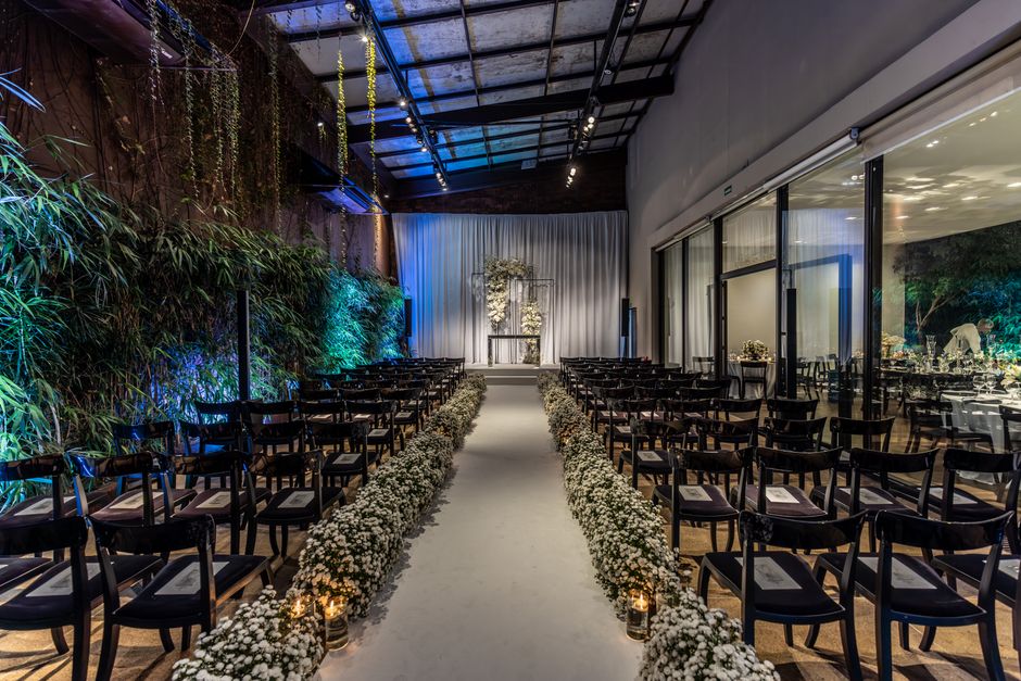 An indoor wedding ceremony setup featuring a central aisle lined with small white floral arrangements in glass jars, leading to a white floral arch on a stage at the front. The aisle is carpeted in wh